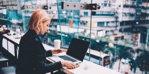 Woman sitting at a table, in a building facing a city view. Coffee sitting on table. She is typing her credit card number into her laptop for a purchase
