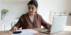 Young, Dark-haired woman, at desk with laptop while typing on calculator 